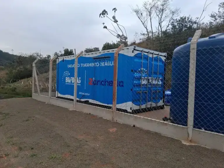 Unidade compacta de tratamento de água instalada ao ar livre, composta por módulos azuis em contêiner cercados por alambrado, com reservatórios plásticos ao lado e área rural ao fundo.