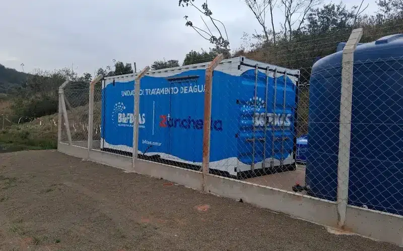 Unidade compacta de tratamento de água instalada ao ar livre, composta por módulos azuis em contêiner cercados por alambrado, com reservatórios plásticos ao lado e área rural ao fundo.